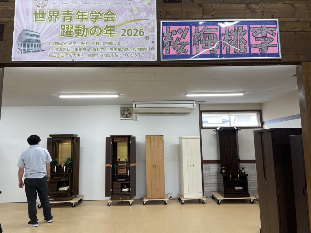 Man in a light blue shirt stands with his back to the camera, inspecting row of wooden Buddhist altars on wheeled platforms in a showroom under pink banners.