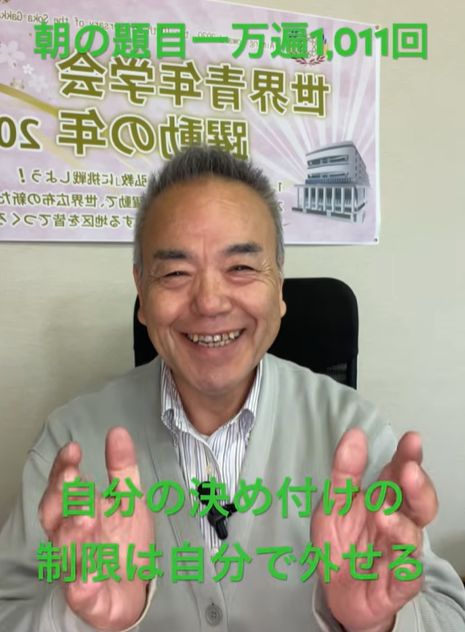 Smiling middle-aged man in a light cardigan sits facing the camera with hands raised; a poster with Japanese text is on the wall behind him, and green captions read '朝のテーマ一万遍1,011回' (top) and '自分の決め付けの制限は自分で外せる' (bottom).