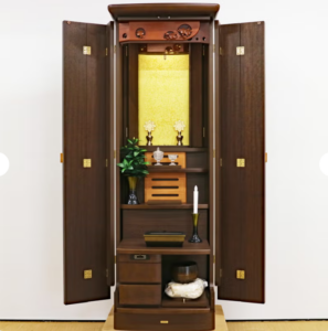 Tall wooden Buddhist home altar (butsudan) opened to reveal shrine with gold backdrop, flowers, candle, and offerings inside a dark cabinet.