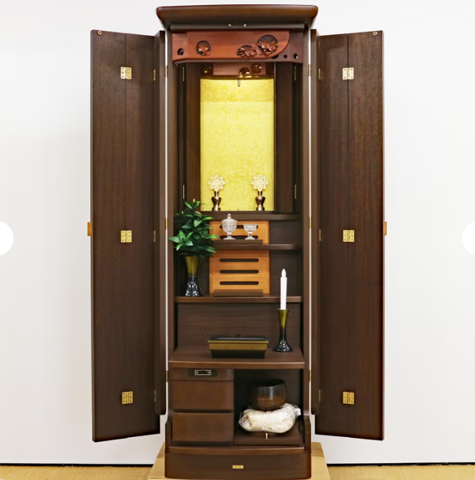 Tall wooden Buddhist home altar (butsudan) opened to reveal shrine with gold backdrop, flowers, candle, and offerings inside a dark cabinet.