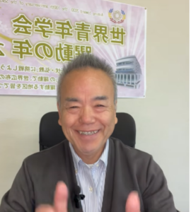 Smiling middle-aged man in a gray jacket sits in an office chair with a purple banner containing Japanese text and a building graphic behind him.
