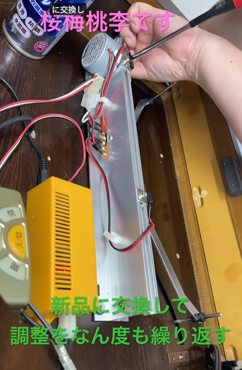 Hands using a screwdriver to adjust wiring inside a clear plastic electronics enclosure with red/black wires and a yellow power module nearby, indicating a repair/assembly scene.