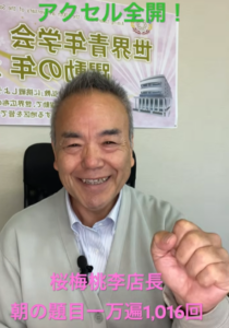 Smiling man in a light gray cardigan recording a video; a poster with Japanese text and a building illustration is behind him with pink captions overlaid.