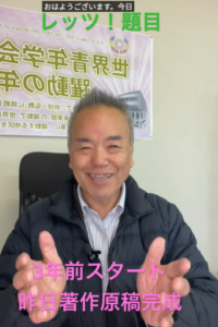 Smiling Japanese man in a dark jacket gestures toward the camera in a classroom, with a banner behind him and pink captions at the bottom saying '3年前スタート 昨日著作原稿完成'.