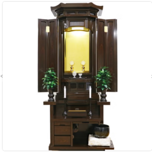 Wooden home Buddhist altar (butsudan) with open doors and a gold-inlaid shrine, candles and small offerings on the center shelf, and potted plants on either side.