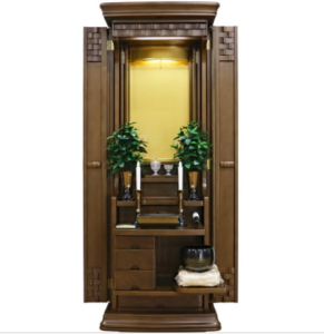 Wooden home shrine/altar cabinet with two potted plants, small offerings, and drawers, against a lit inner alcove.
