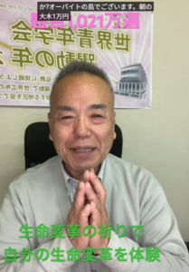 Smiling man in a gray jacket sits with hands pressed together in a prayer-like gesture, in front of a poster with Japanese text and a drawing of a building; green caption at bottom reads about life transformation.