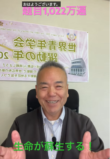 Smiling middle-aged man in a dark jacket seated in an office, with a pink poster featuring Japanese text and a building image behind him; top caption reads 'おはようございます' and green caption at bottom says '生命が蘇生する！'