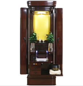 Open dark wood Buddhist altar cabinet with illuminated inner shrine, small plants, and prayer items arranged on a central altar.