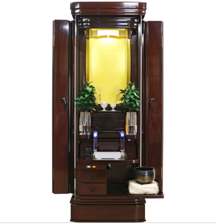 Open dark wood Buddhist altar cabinet with illuminated inner shrine, small plants, and prayer items arranged on a central altar.
