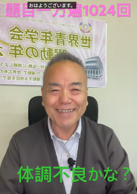 Smiling man seated in a chair with a banner behind him; green text at the bottom reads '体調不良かな？' and a small caption at the top says 'おはようございます'.