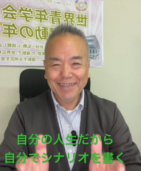 Smiling man in a blazer delivering a message in an office; a banner with Japanese text is on the wall behind him, and green caption text at the bottom reads '自分の人生だから 自分でシナリオを書く'.