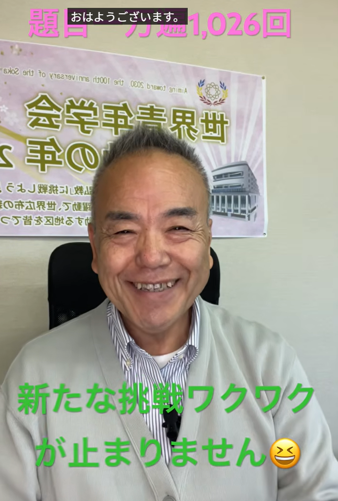 Smiling Asian man in a light gray jacket sits in an office, a banner with Japanese text behind him, and bold pink top text and a green caption at the bottom reading '新たな挑戦ワクワクが止まりません' with a grinning emoji.