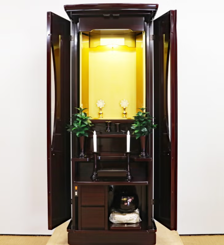 Open dark wooden altar cabinet with a gold interior, featuring candles, small floral ornaments, and potted plants on tiered shelves.