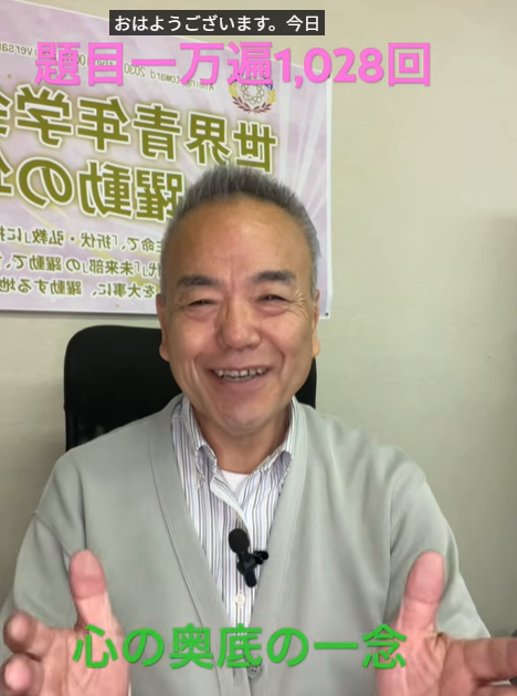Smiling man in a light cardigan sitting in an office, giving two thumbs up; poster with Japanese text behind him and green subtitle '心の奥底の一念' at the bottom.