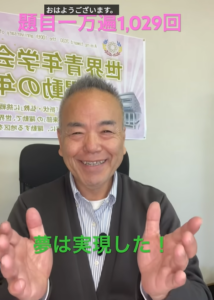 笑顔の男性が両手を広げて挨拶する場面。背景に横断幕と“夢は実現した！”の緑文字テキストがある。