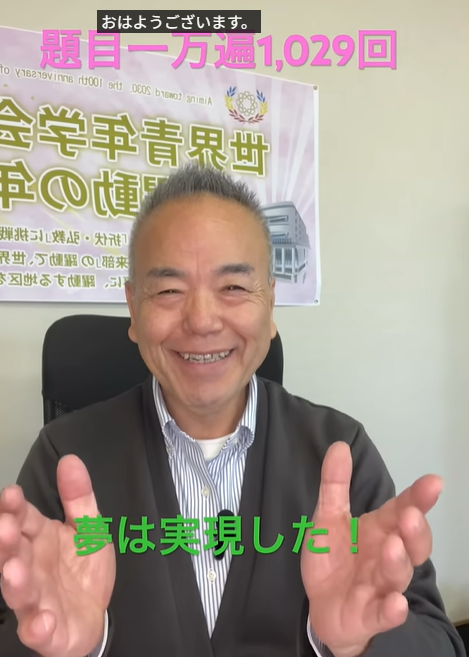 笑顔の男性が両手を広げて挨拶する場面。背景に横断幕と“夢は実現した！”の緑文字テキストがある。