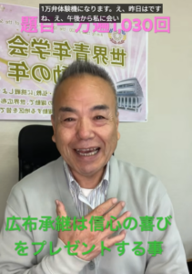 Smiling man in a light gray cardigan sits in an office; green caption at the bottom reads '広布承継は信心の喜びをプレゼントする事'.