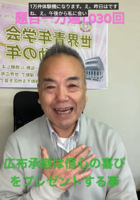 Smiling man in a light gray cardigan sits in an office; green caption at the bottom reads '広布承継は信心の喜びをプレゼントする事'.