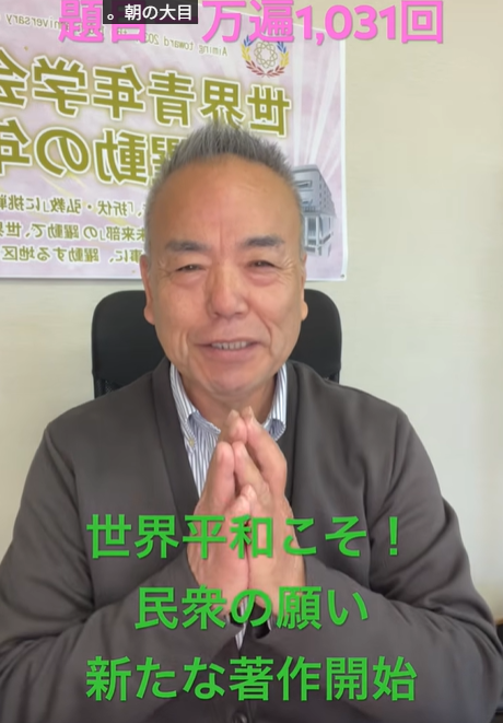 Older man in a brown cardigan sits in an office, hands pressed together in a prayer gesture, smiling at the camera; a banner with Japanese text is on the wall behind him. Green text overlay reads: 'World peace is the goal! The people's wish; start of a new work'