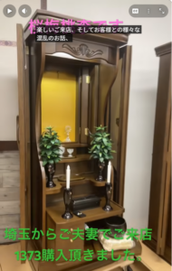 Small wooden home altar with two candlesticks and potted plants in front of a yellow back panel, inside a carved cabinet.