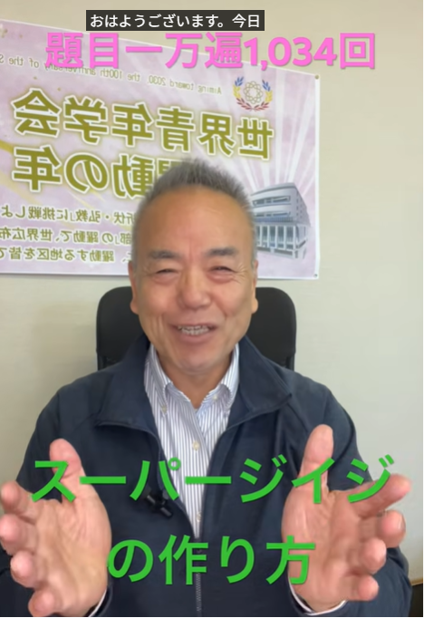 Smiling Japanese man seated indoors, gesturing with both hands as if explaining, with a poster and bold green subtitle text in Japanese at the bottom of the screen.