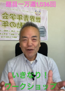 Smiling Japanese man in a striped shirt sits in an office, making a peace sign with both hands, with a large Japanese poster on the wall behind him.