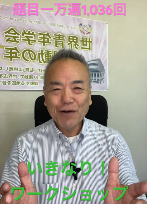 Smiling Japanese man in a striped shirt sits in an office, making a peace sign with both hands, with a large Japanese poster on the wall behind him.