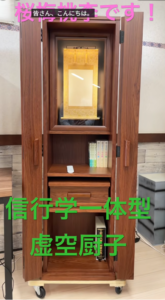 Wooden butsudan on wheels with an inner gold shrine, open doors, and a few books on a shelf; pink banner text at top and green banner text across the front in Japanese.