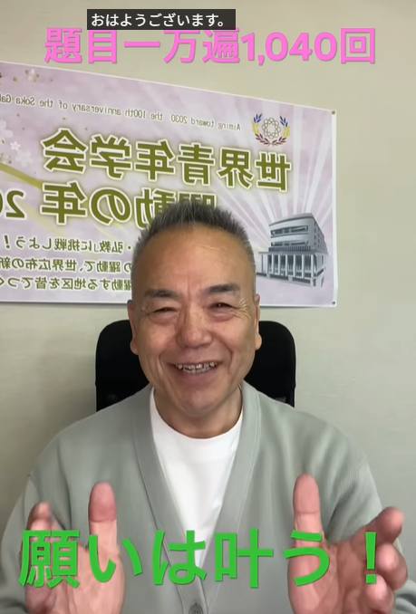 Smiling Japanese man seated in a chair, hands raised, with a banner in the background and green caption '願いは叶う！' at the bottom; a small black 'おはようございます。' caption at the top.