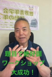 Smiling man in a blue polo speaking to the camera during a workshop, with a banner in the background and green Japanese caption claiming the workshop was a great success.