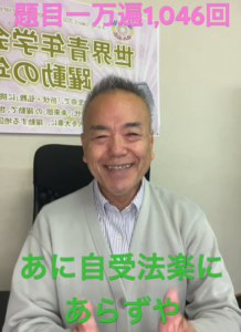 Smiling man in a light cardigan sits in an office chair, with a poster behind him and pink top caption plus green bottom caption in Japanese.