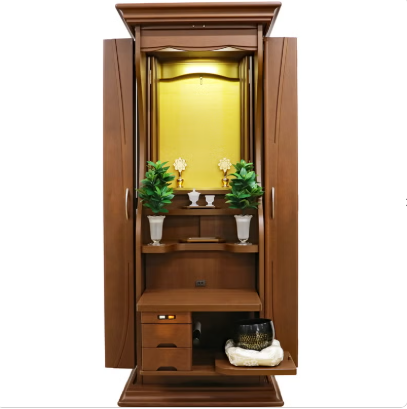 Open wooden home altar (butsudan) with a yellow back panel, two small potted plants, candles, and shelves for offerings.