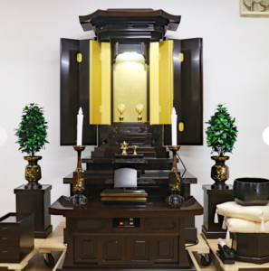 Butsudan-style household altar: dark wood cabinet with gold inner doors open, central shrine and small devotional objects, flanked by two white-candle holders on either side and green potted plants.