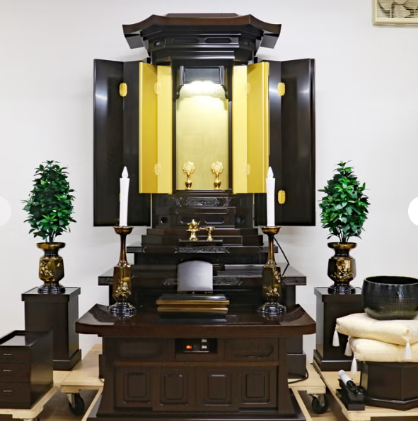 Butsudan-style household altar: dark wood cabinet with gold inner doors open, central shrine and small devotional objects, flanked by two white-candle holders on either side and green potted plants.