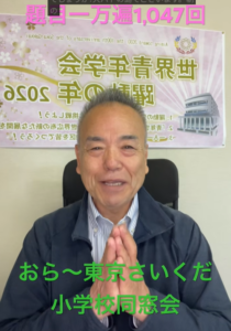 Smiling older man seated in an office, hands pressed together in a greeting, with a poster behind him.