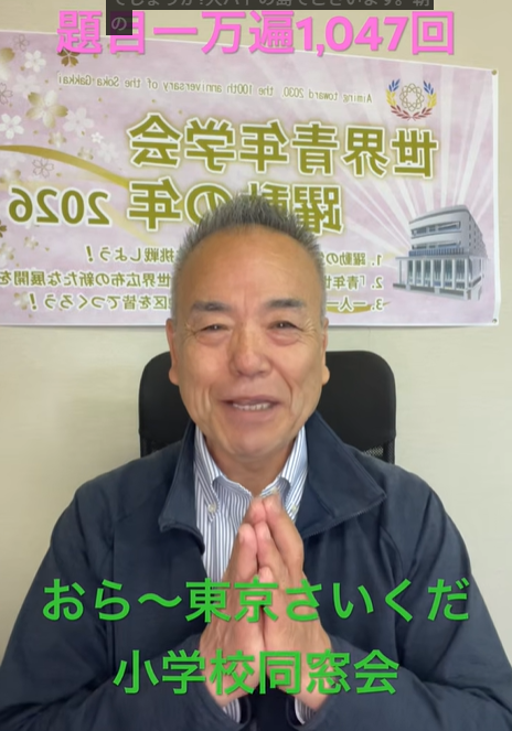 Smiling older man seated in an office, hands pressed together in a greeting, with a poster behind him.