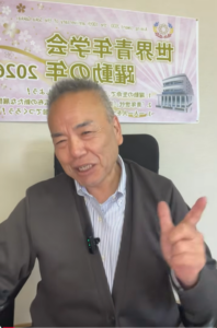 Smiling man in a blazer seated indoors, giving a peace sign; a purple banner with Japanese text and a building graphic is behind him.
