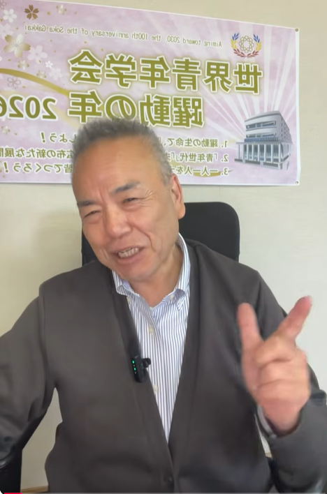 Smiling man in a blazer seated indoors, giving a peace sign; a purple banner with Japanese text and a building graphic is behind him.