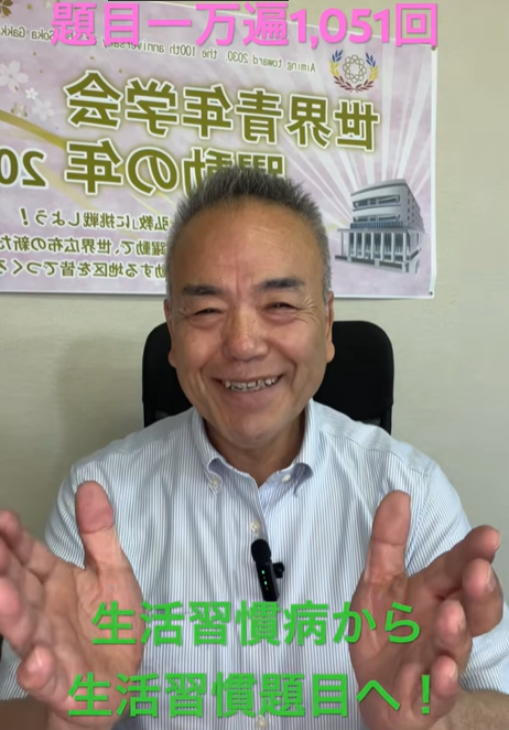 Smiling man in a striped shirt sits in an office, hands raised toward the camera; a banner/poster and green Japanese captions appear behind him.