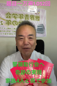 Smiling middle-aged man in a white shirt sits in an office, holding several red booklets with a banner in Japanese behind him.
