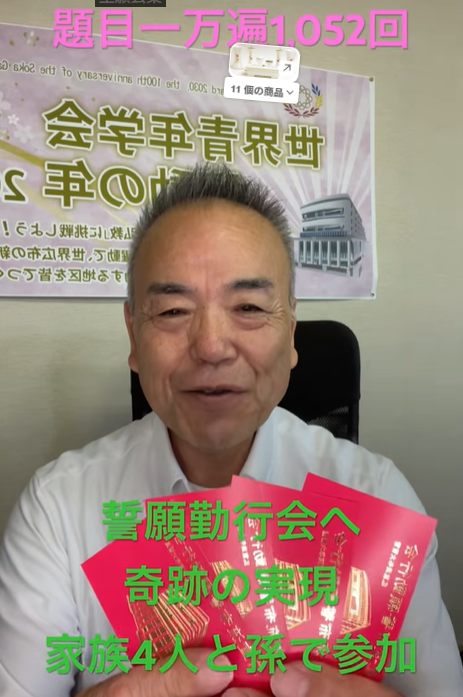 Smiling middle-aged man in a white shirt sits in an office, holding several red booklets with a banner in Japanese behind him.