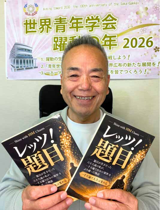 Smiling man seated at a desk holds two black-and-gold Japanese books titled 'レッツ! 問題!' in front of a pink banner with 2026 text in the background.