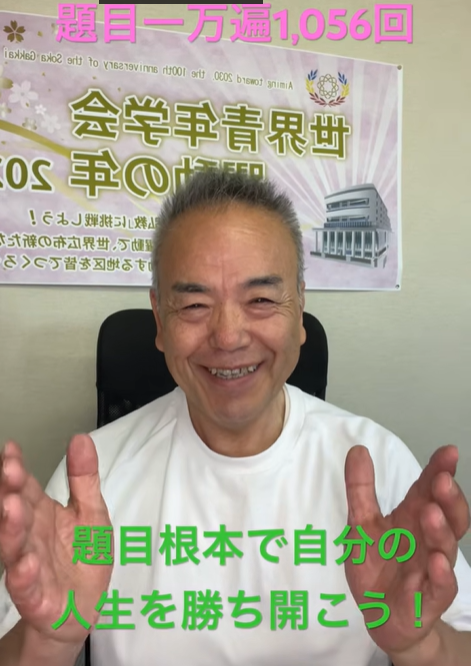 Smiling middle-aged man in a white shirt sits in an office chair, hands raised toward the camera; a poster with Japanese text and a building image is on the wall behind him; green Japanese caption at the bottom reads '題目根本で自分の人生を勝ち開こう！' and pink caption at the top says '題目一万遍1,056回'