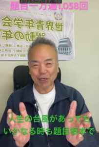 Smiling man in a dark jacket sits in front of a poster, with pink caption at top and green caption at bottom in Japanese text.