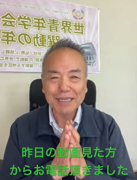 Smiling older man seated in a chair, hands pressed together in a greeting gesture, with a banner of Japanese text behind him.