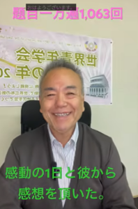 Smiling middle‑aged man in a suit sits in an office chair with a banner in the background and green Japanese captions at the bottom that read 感動の1日と彼から感想を頂いた。