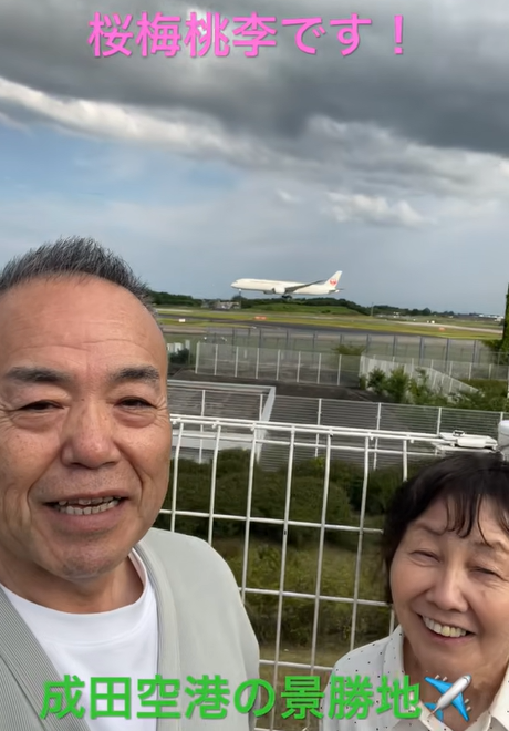 Smiling couple taking a selfie at Narita Airport as a plane lifts off in the background; pink top text 桜梅桃李です！, green bottom text 成田空港の景勝地.
