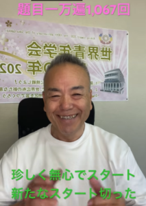 Smiling man in a white shirt sits in front of a banner with Japanese text in a room setting.