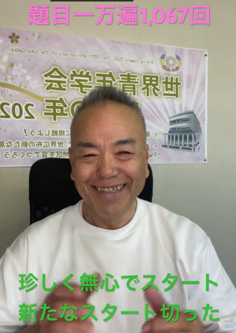 Smiling man in a white shirt sits in front of a banner with Japanese text in a room setting.